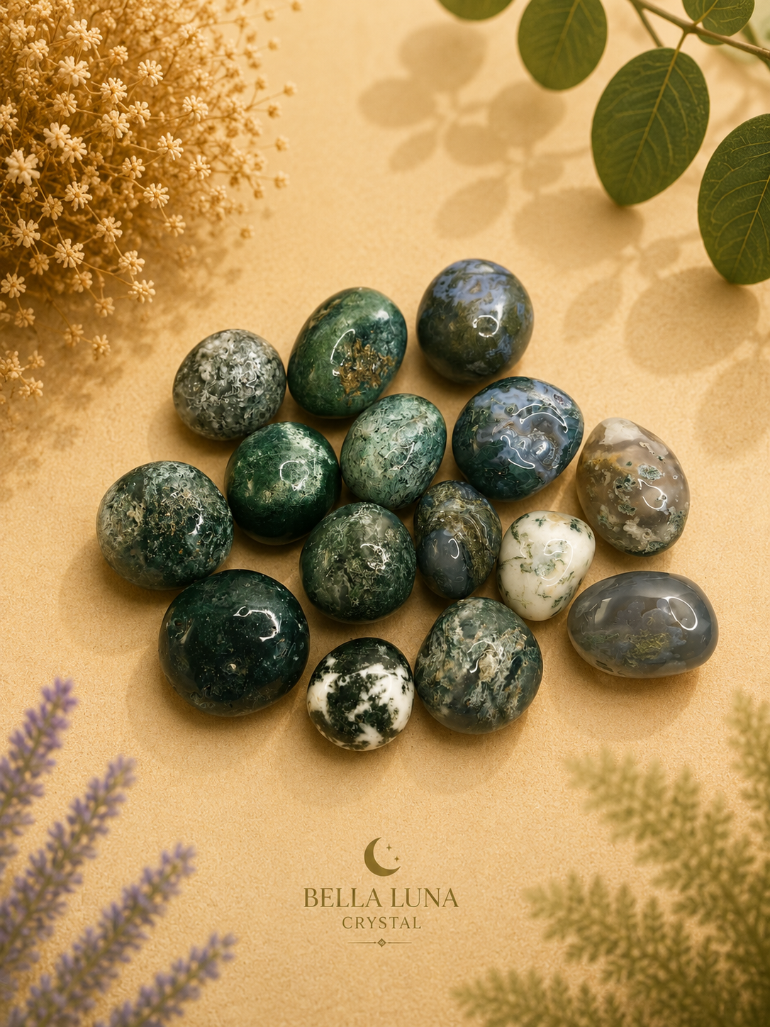 A cluster of polished green and blue jasper stones arranged on a beige surface with dried flowers and green leaves around them. The brand name Bella Luna Crystal is printed at the bottom.