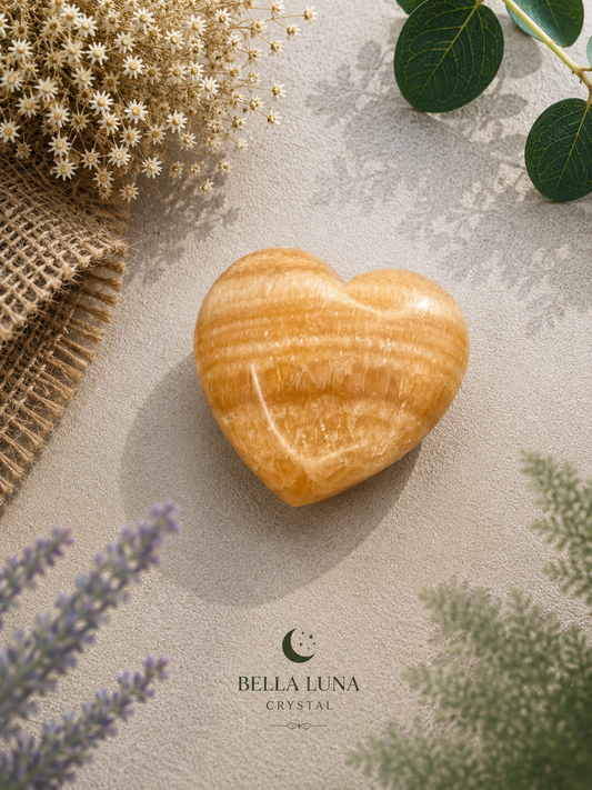 A polished heart-shaped crystal with warm golden-orange and brown banding, displayed on a textured beige surface with dried flowers, green leaves, and lavender sprigs. The brand logo 'BELLA LUNA CRYSTAL' is visible at the bottom.