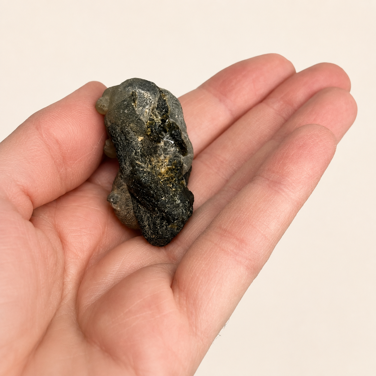 A rough, dark gray metallic mineral specimen with gold flecks, held in the palm of a hand against a plain light beige background.