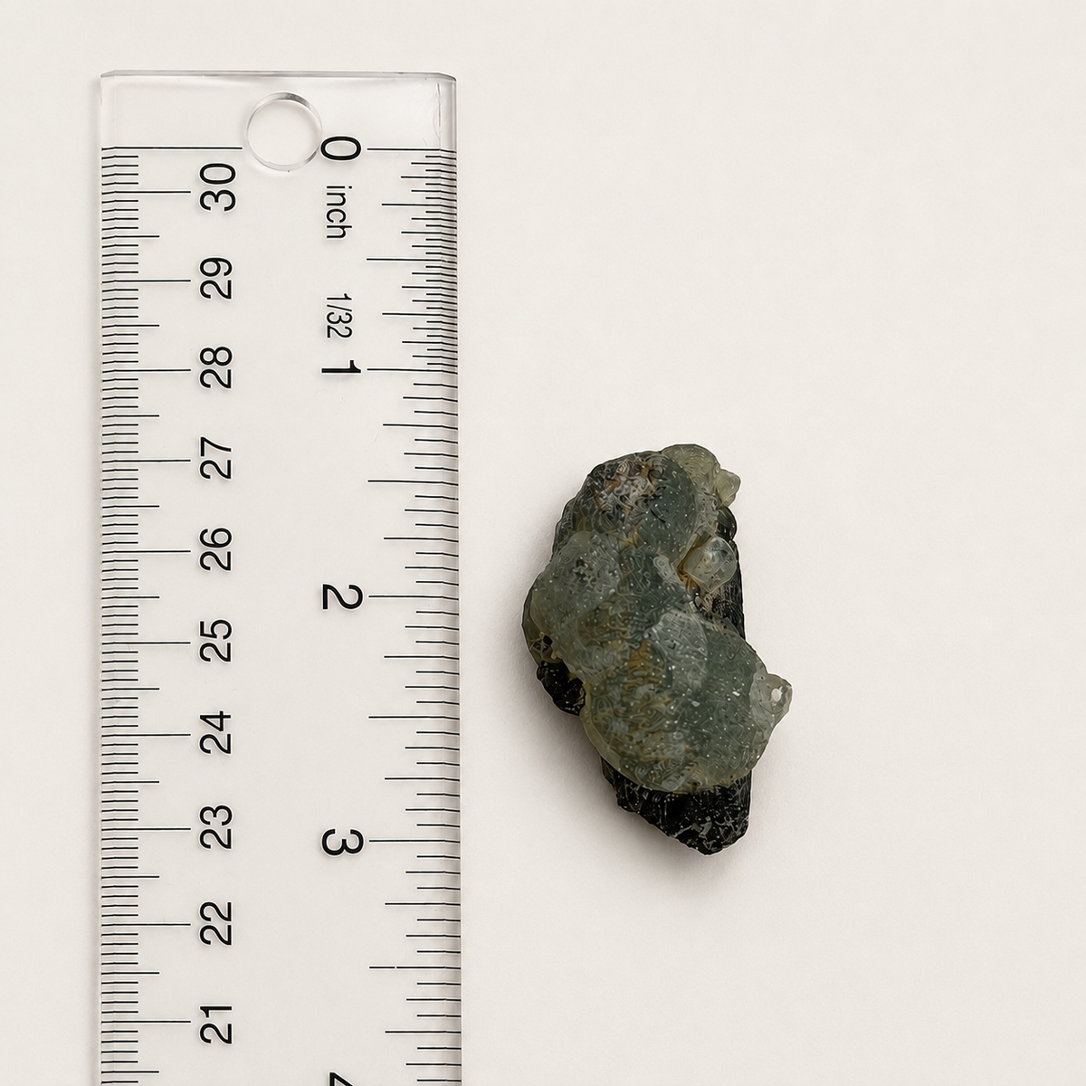 A small rough green stone or mineral sample placed beside a clear plastic ruler with inch markings on a plain light background.