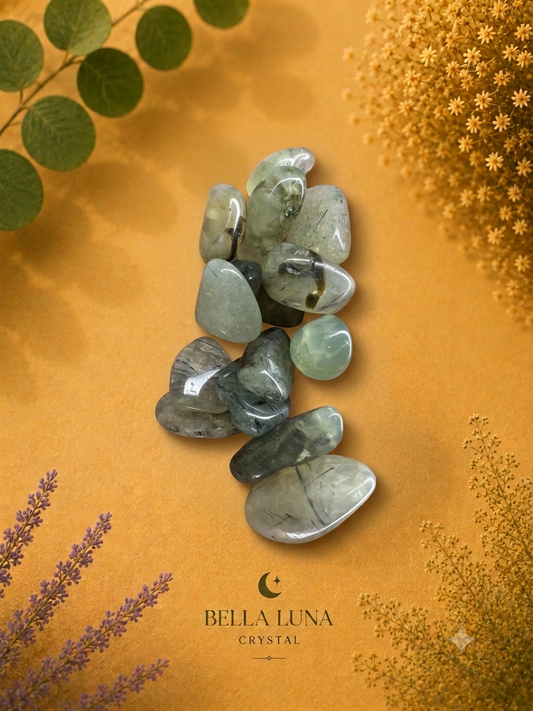 A cluster of polished tumbled crystals in pale green, gray, and smoky tones arranged on a warm mustard-yellow background with decorative dried flowers and greenery. The brand name Bella Luna Crystal is printed at the bottom.