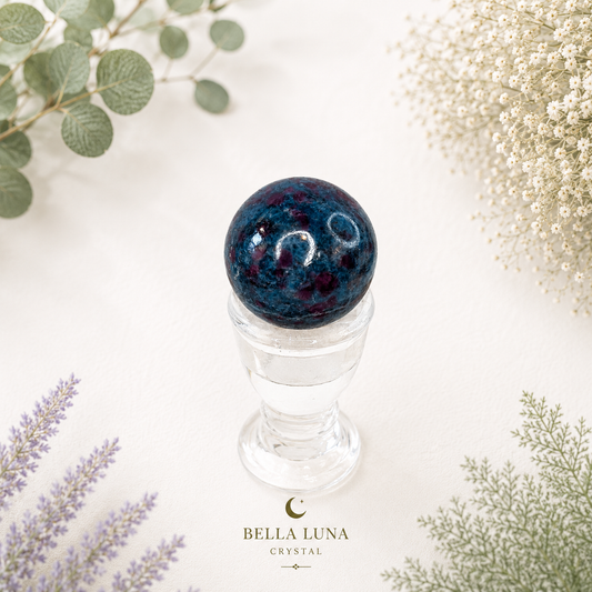 A dark teal and red kyanite crystal ball resting in a clear glass holder, styled with dried flowers and greenery on a white background. The text 'BELLA LUNA CRYSTAL' is visible at the bottom.