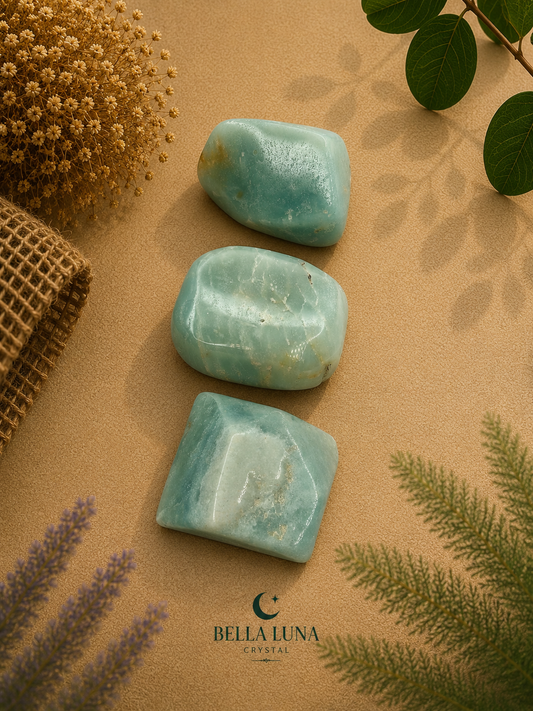 Three polished light blue-green crystal stones arranged vertically on a beige textured surface with dried flowers, green leaves, and a folded burlap fabric. The Bella Luna Crystal logo appears at the bottom.