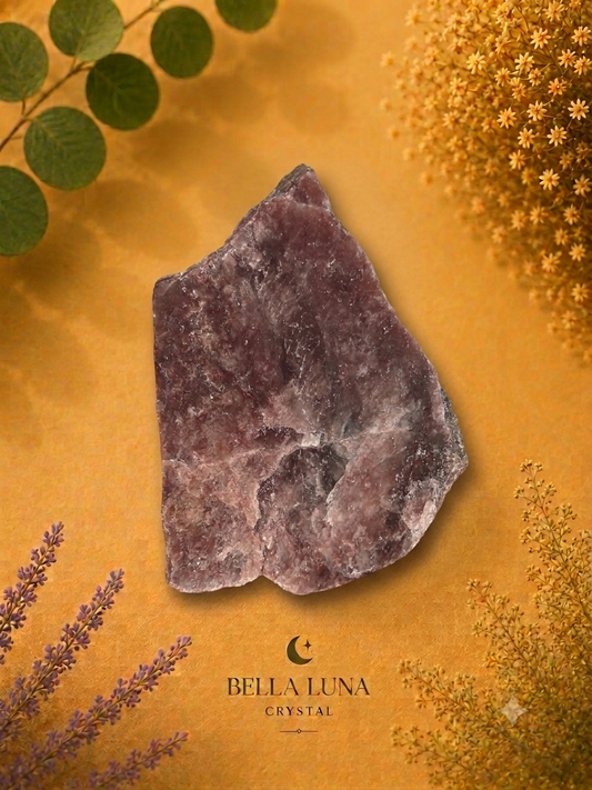 A rough, unpolished purple crystal stone with natural facets, displayed on a textured mustard yellow background decorated with dried botanicals and green leaves. The brand logo 'BELLA LUNA CRYSTAL' is visible at the bottom.