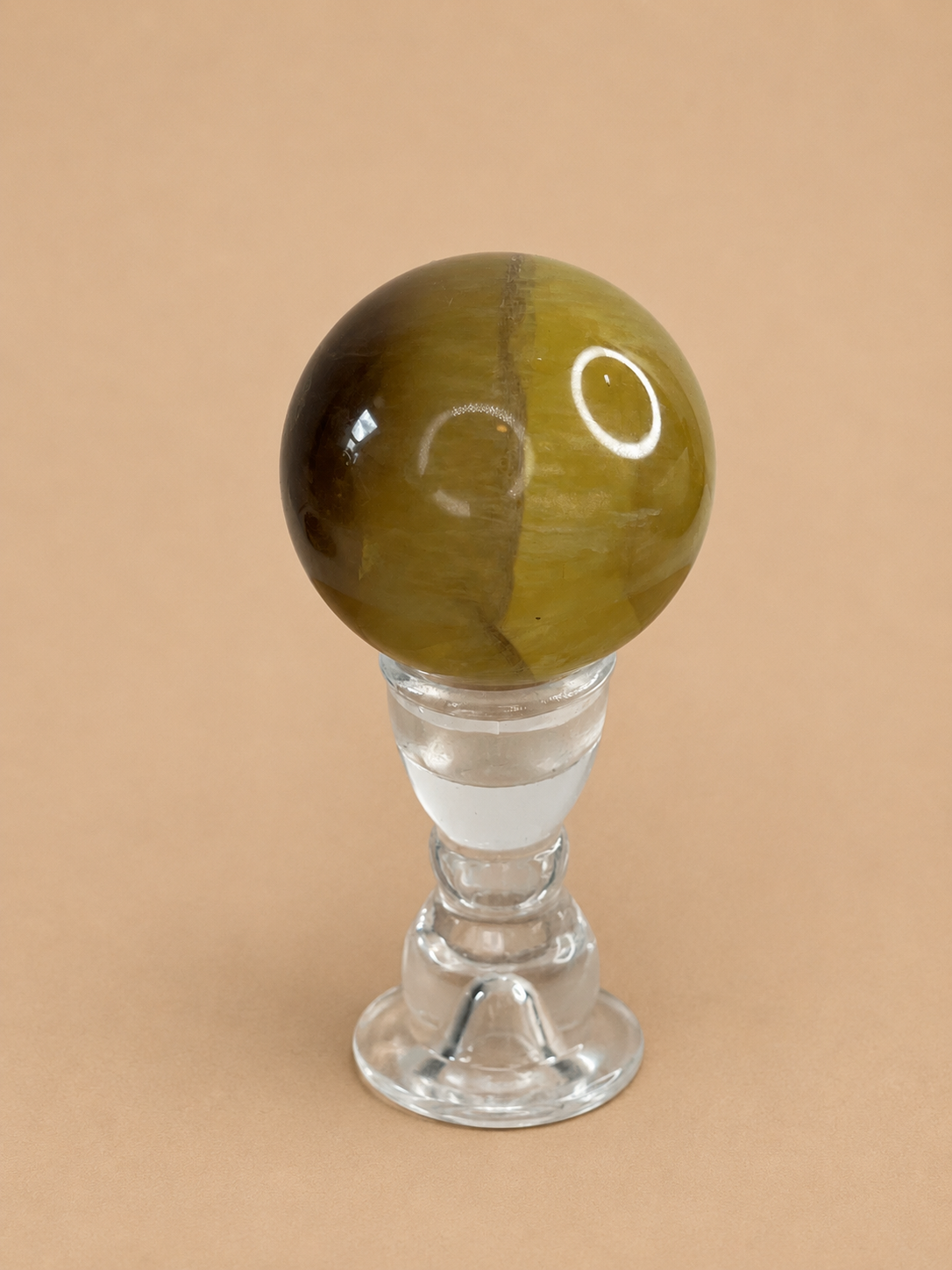 A polished fluorite sphere on a clear glass stand, set against a solid beige background.