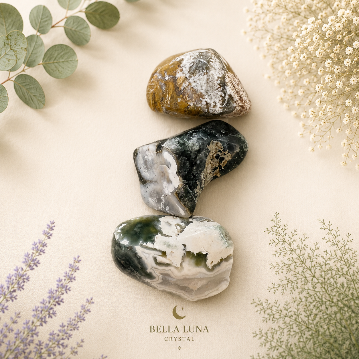 Three polished tumbled crystals stacked vertically on a beige surface with botanical props. The crystals have natural marbled patterns in green, white, gray, and brown.