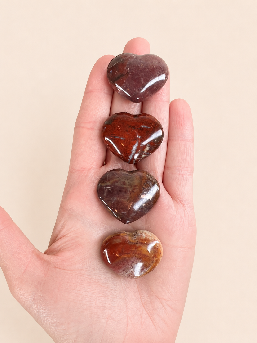 Four polished heart-shaped stone pendants in shades of brown and reddish-brown, held vertically in a person's open hand against a plain beige background.
