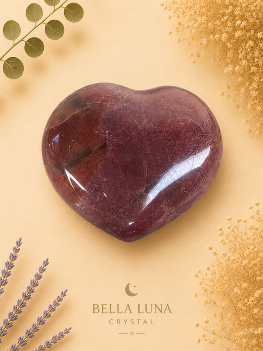 A polished heart-shaped crystal stone in deep reddish-brown tones, displayed on a warm beige background with decorative dried flowers and greenery. The brand name 'BELLA LUNA CRYSTAL' is printed at the bottom.