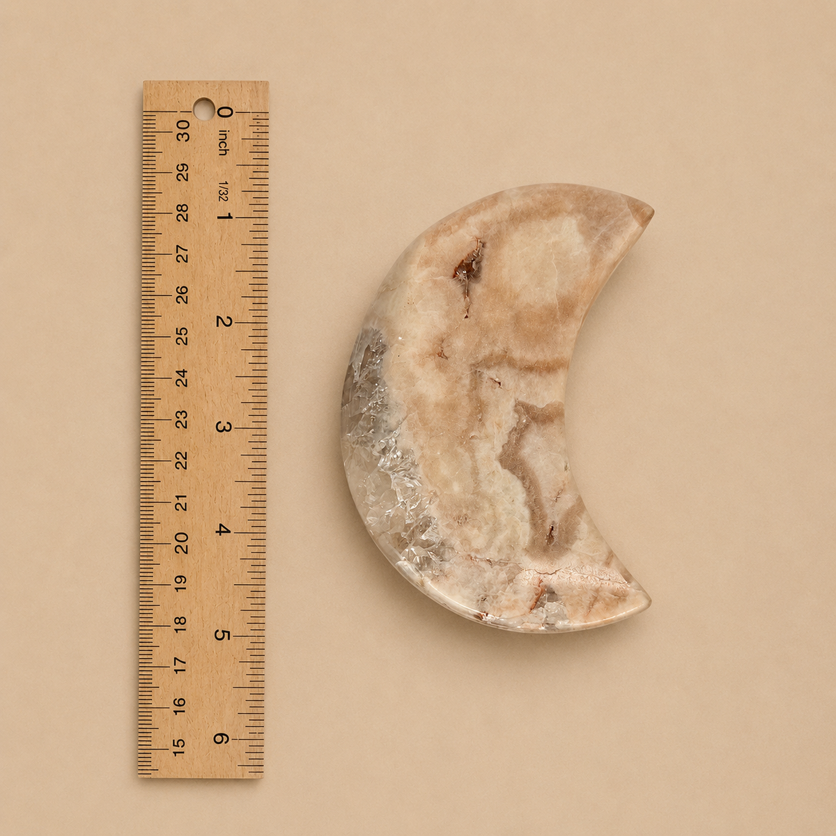 A crescent-shaped polished stone object with natural beige, cream, and light brown marbling, shown next to a wooden ruler on a plain beige background.