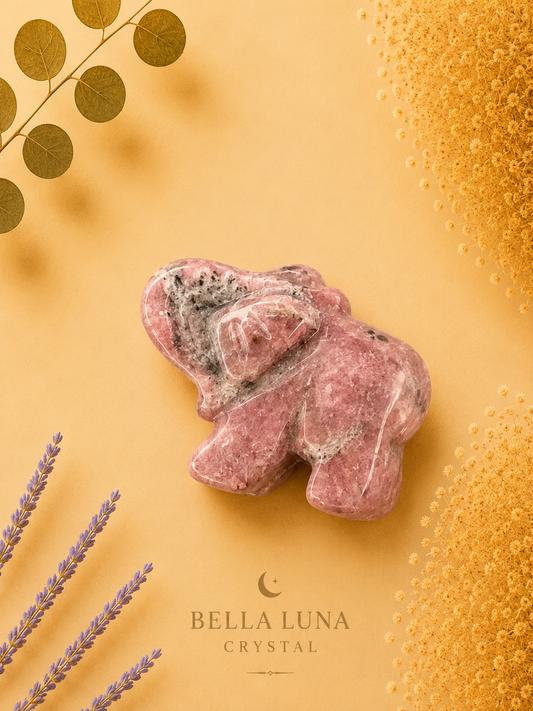A pinkish crystal elephant figurine with natural marbling, styled on a warm yellow background with decorative gold leaves, lavender sprigs, and a textured gold pattern. The brand name 'BELLA LUNA CRYSTAL' is printed at the bottom.