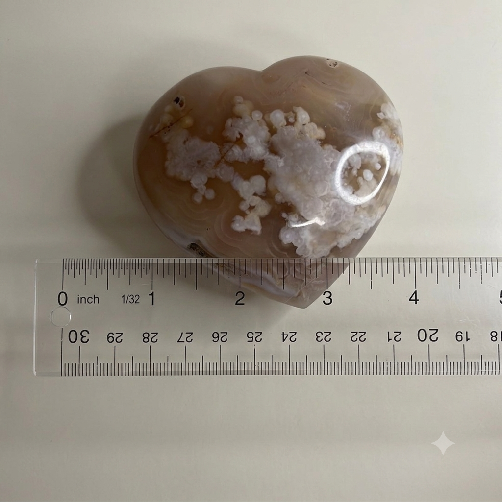 A heart-shaped agate stone with natural white and pale pink crystalline patterns, shown next to a clear plastic ruler for scale on a light beige background.