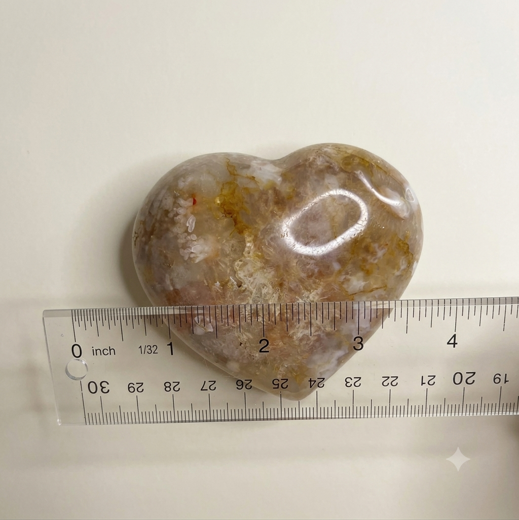 A polished heart-shaped stone with natural brown, beige, and white marbling, shown next to a clear plastic ruler for size reference on a plain off-white background.