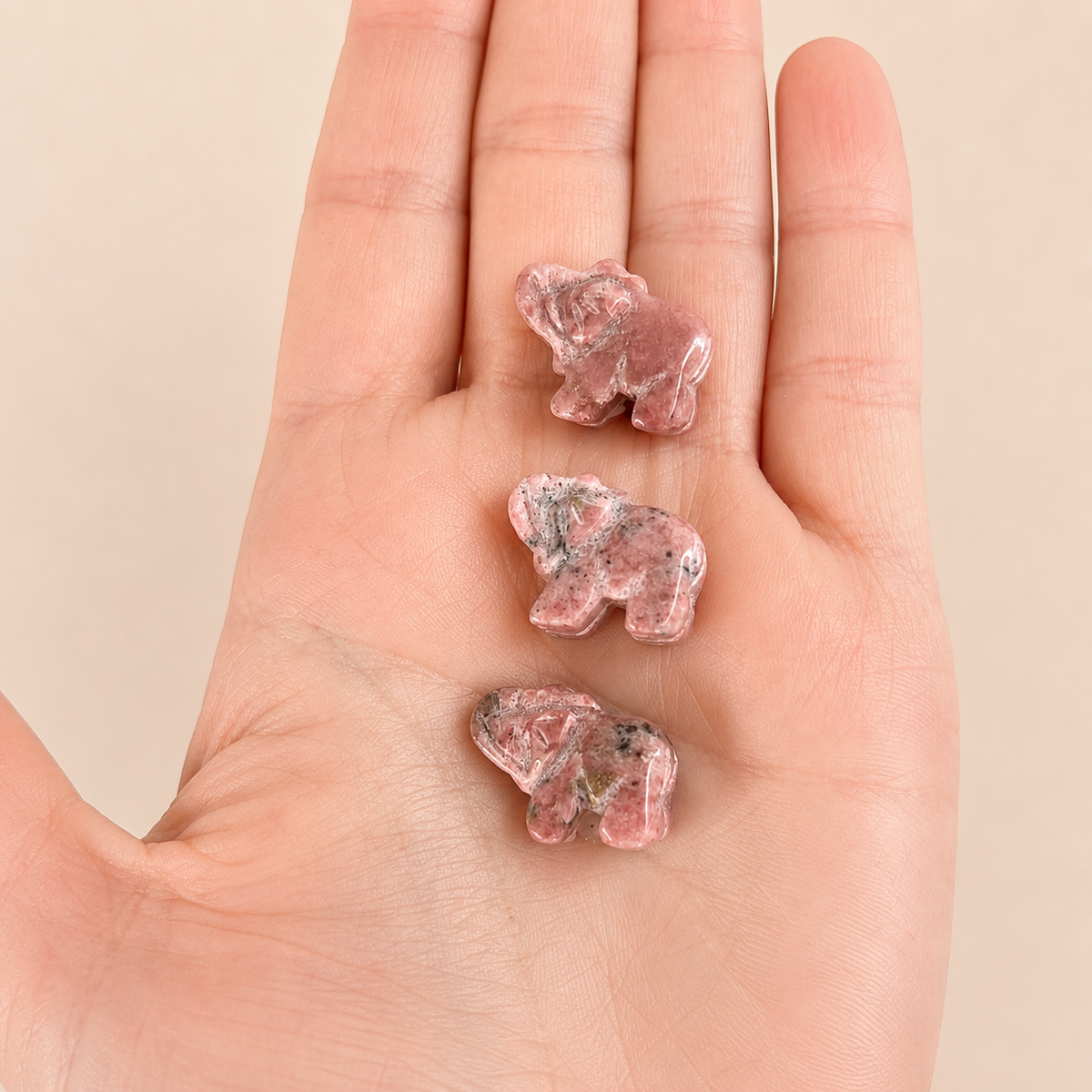Three small pink crystal elephant charms with dark inclusions are displayed on an open human hand against a soft beige background.