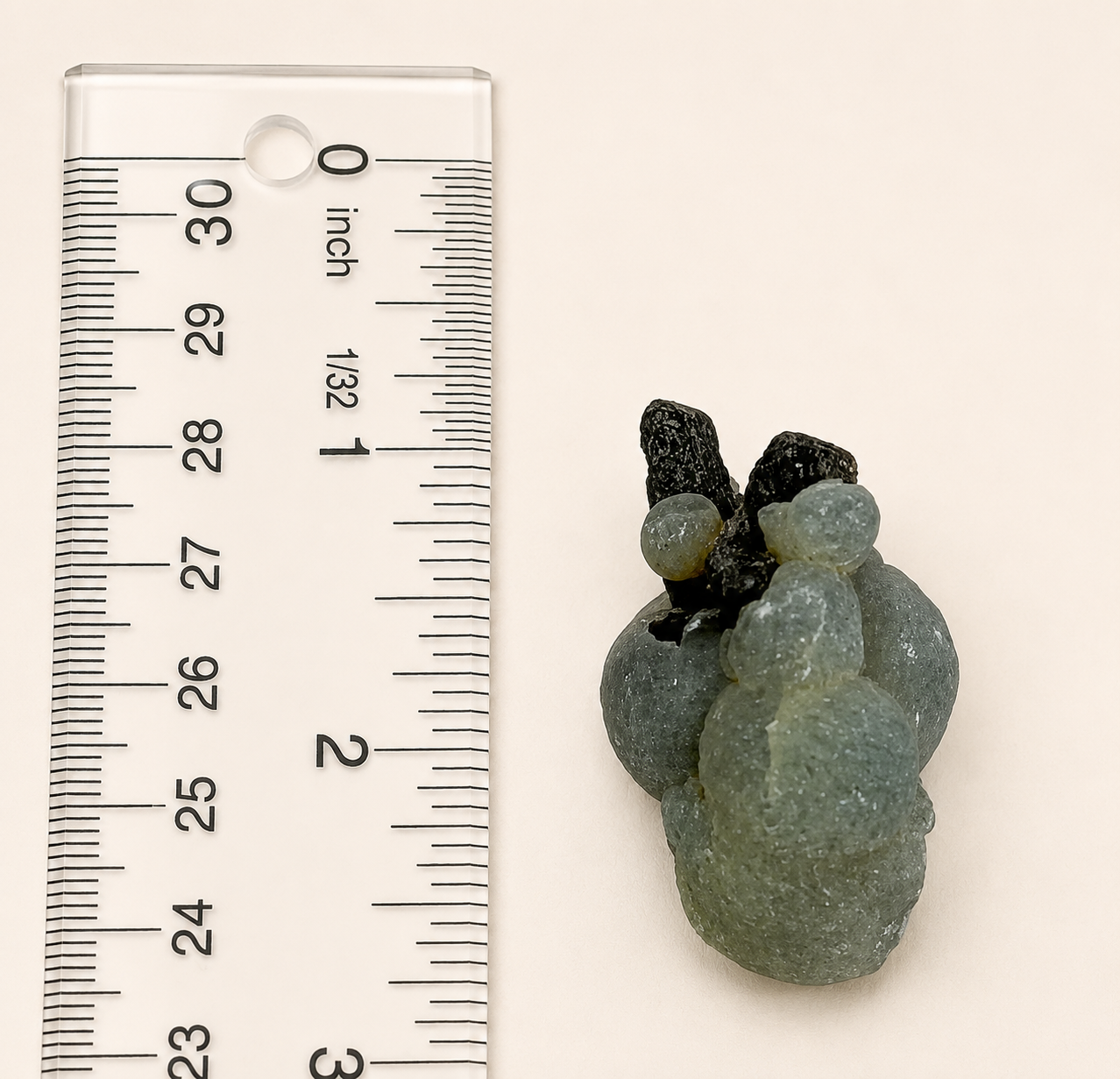 A small carved stone ornament or bead cluster in pale green-gray with dark black accents, shown next to a clear plastic ruler for scale on a plain light background.