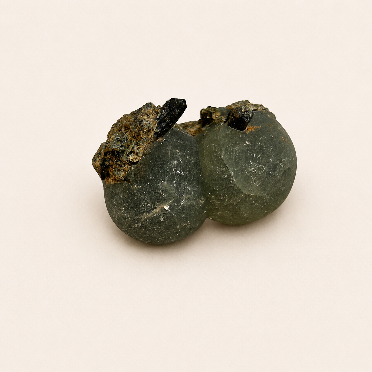 A natural mineral specimen featuring two smooth, rounded dark greenish-gray boulder-like forms fused together, with rough brown and black matrix material on top, photographed on a plain light beige background.