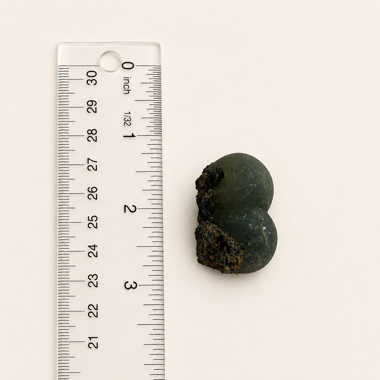 A small dark green, rounded natural specimen with rough brown areas, shown next to a clear plastic ruler with inch markings on a plain off-white background.