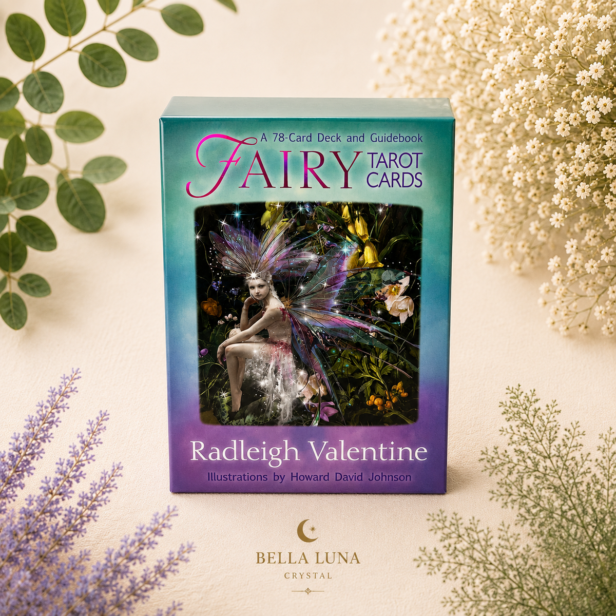 A boxed card deck and guidebook titled 'Fairy Tarot Cards' by Radleigh Valentine, photographed upright on a light cream surface with decorative green and white botanicals around it. The box has a teal-to-purple gradient cover with a fairy illustration in the center.