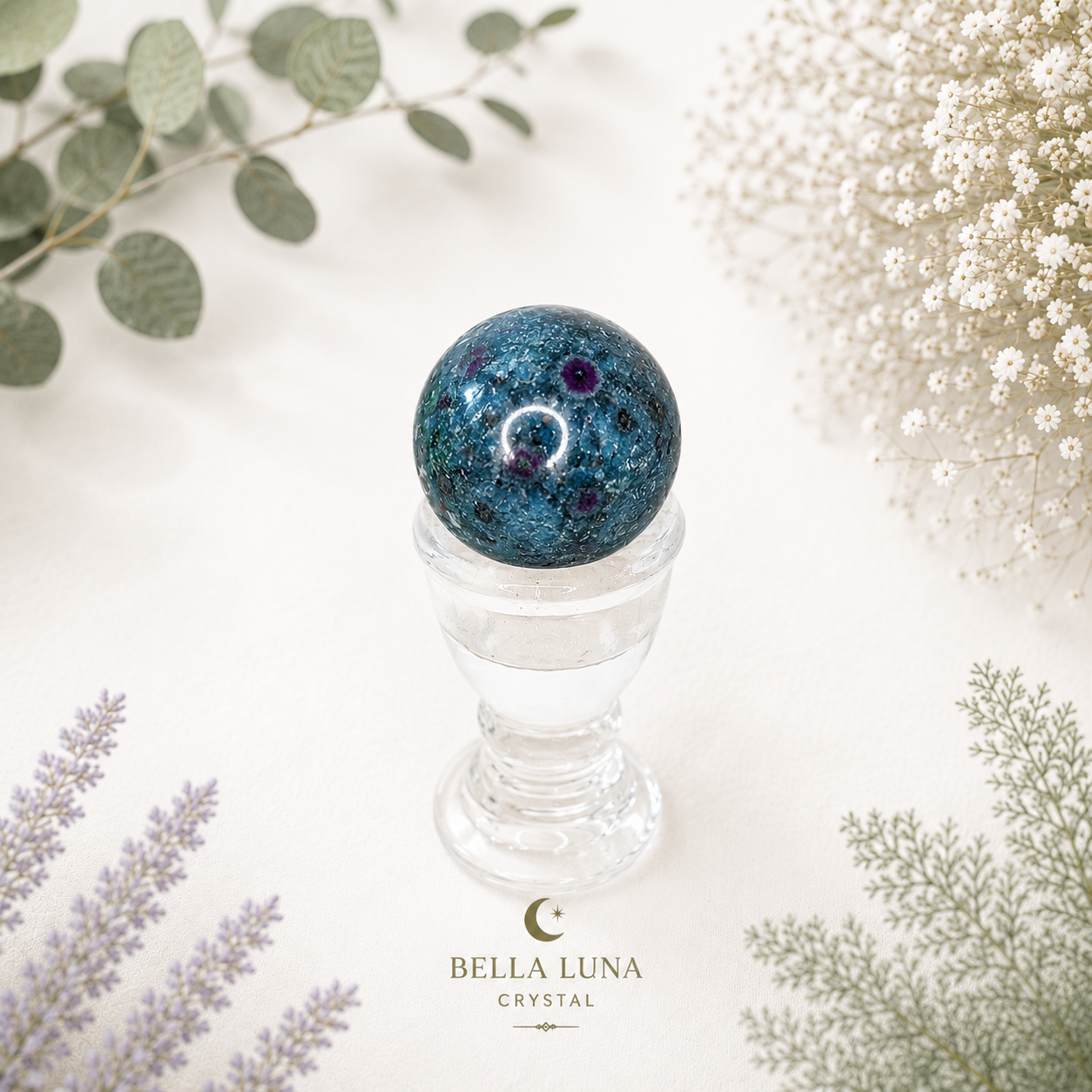 A decorative crystal ball with a deep blue and black starry pattern sits on a clear glass pedestal. The scene is styled with dried white flowers, green leaves, and lavender sprigs on a white background, with the brand name 'BELLA LUNA CRYSTAL' printed at the bottom.
