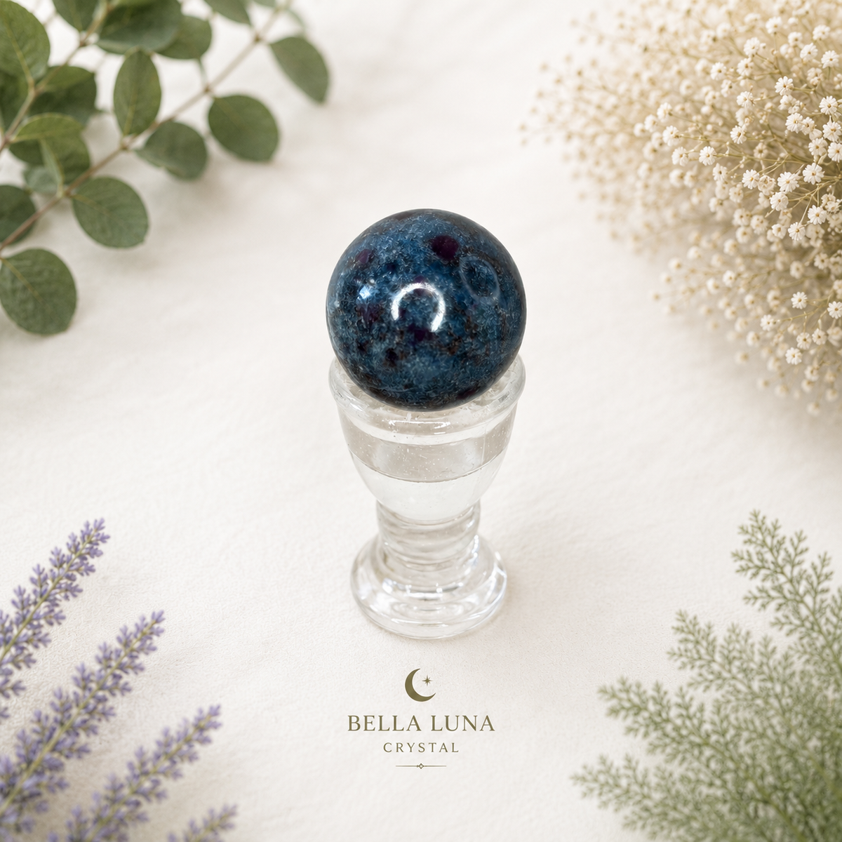 A dark blue speckled crystal ball displayed on a clear glass pedestal, surrounded by dried flowers and green botanical leaves on a light cream background. The brand name 'BELLA LUNA CRYSTAL' is printed at the bottom.