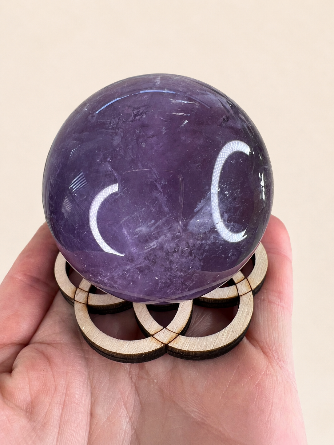 A polished purple amethyst crystal sphere resting on a small wooden stand, held in a person's hand against a plain beige background.