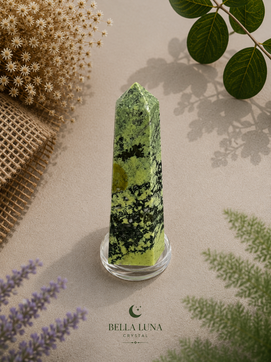 A green and black mottled crystal tower on a clear glass base, styled on a beige surface with dried flowers, burlap, leaves, and lavender sprigs. The brand name 'BELLA LUNA CRYSTAL' is printed at the bottom.