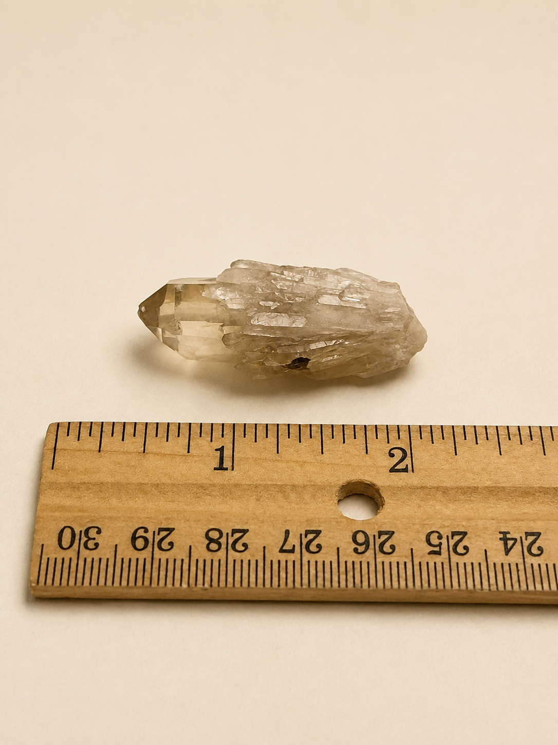 A small natural clear quartz crystal point shown above a wooden ruler for scale on a plain beige background.