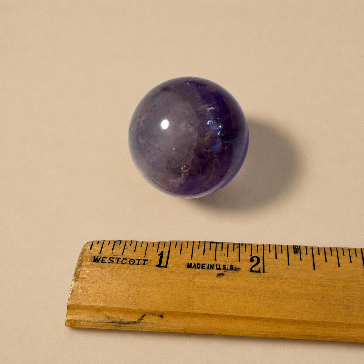 A polished purple amethyst sphere shown above a wooden ruler for scale on a plain beige background.