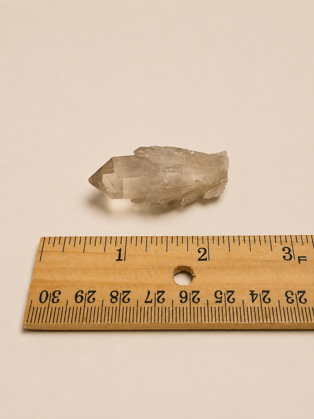 A small rough translucent crystal cluster placed above a wooden ruler on a plain light beige background, with the ruler showing measurements in centimeters and inches.