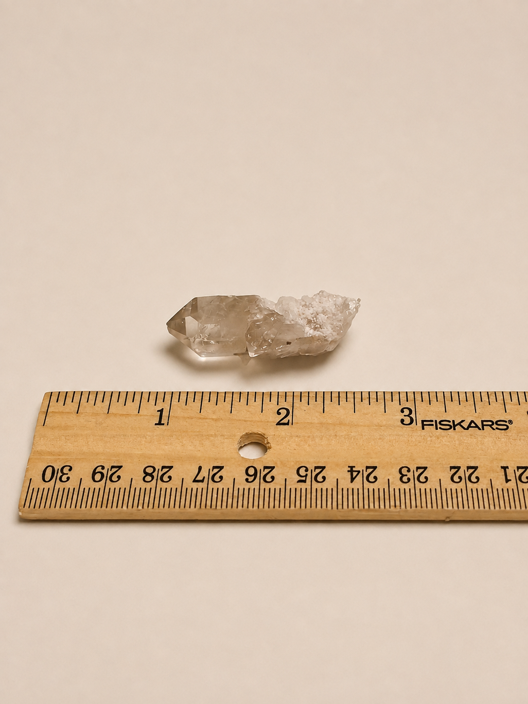 A small natural clear quartz crystal cluster placed above a wooden Fiskars ruler on a plain beige background, showing its size.