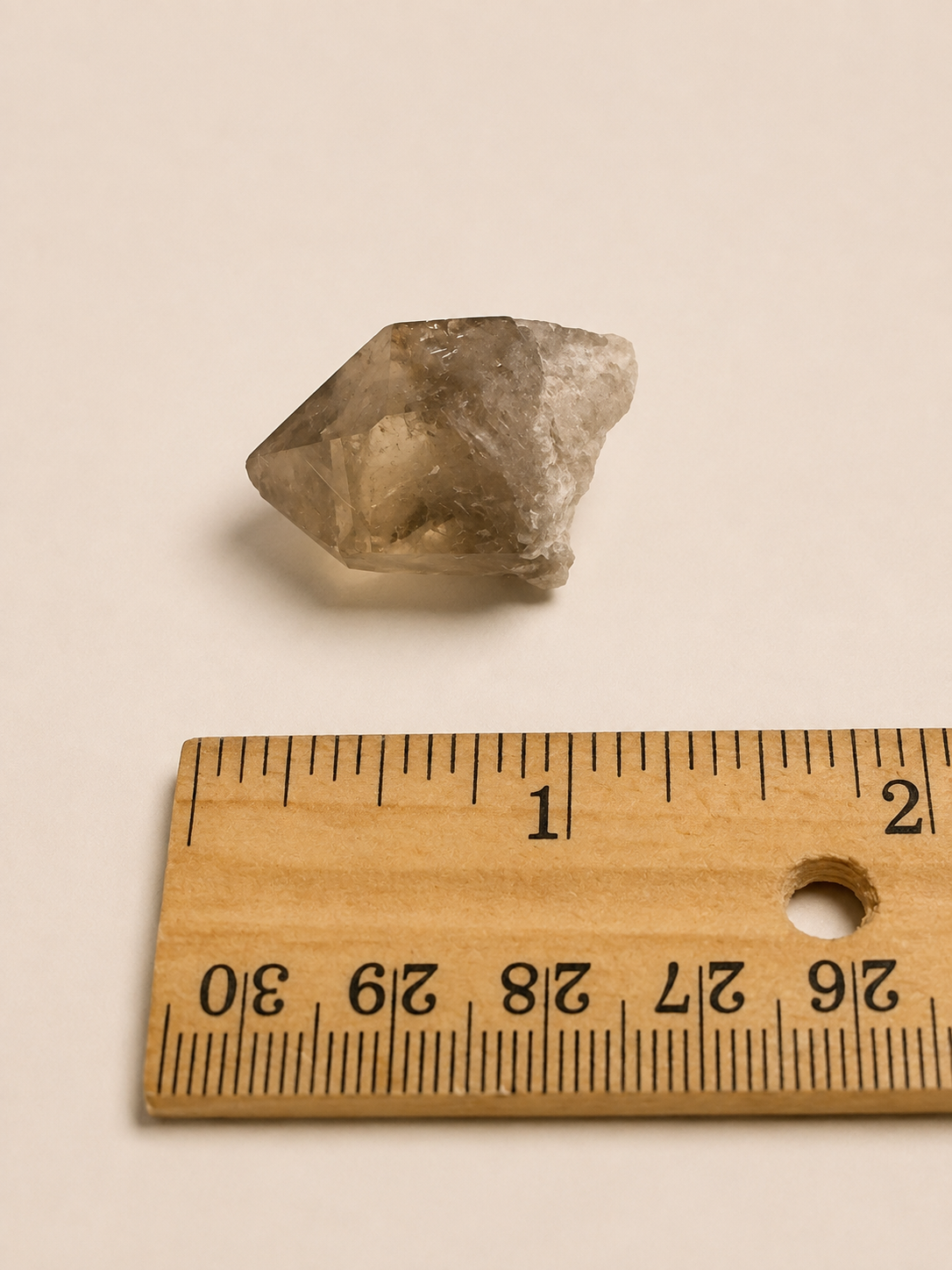 A small translucent gray crystal or stone placed above a wooden ruler on a plain light beige background.