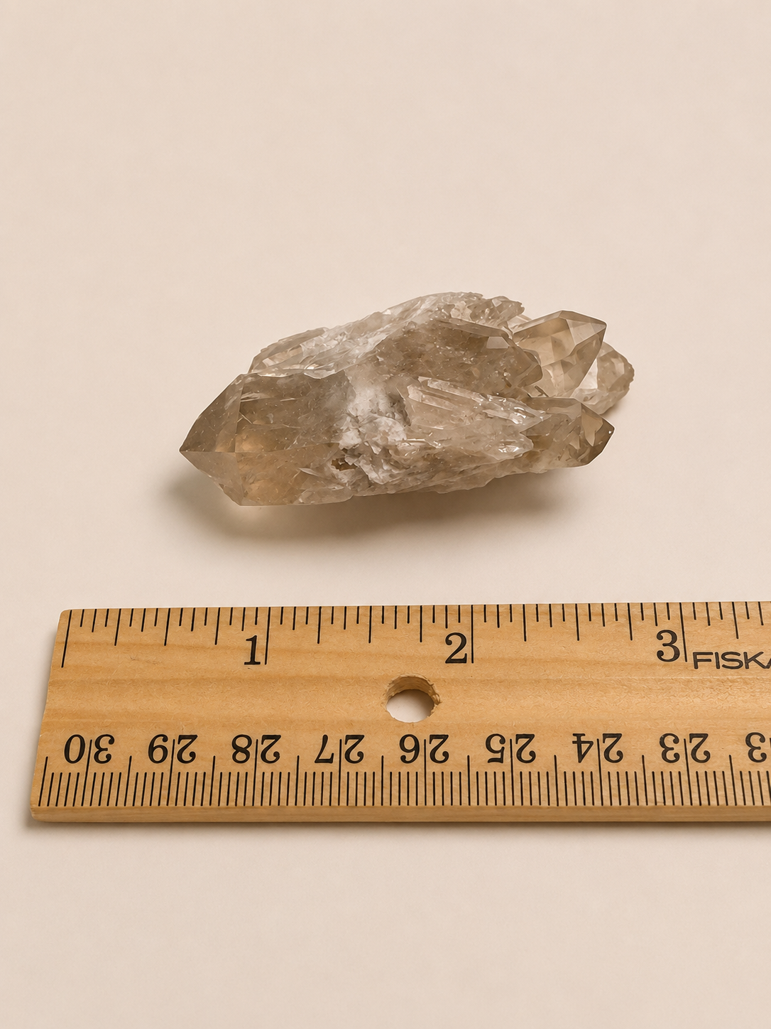 A raw, translucent smoky quartz crystal cluster shown next to a wooden ruler for scale on a plain beige background.