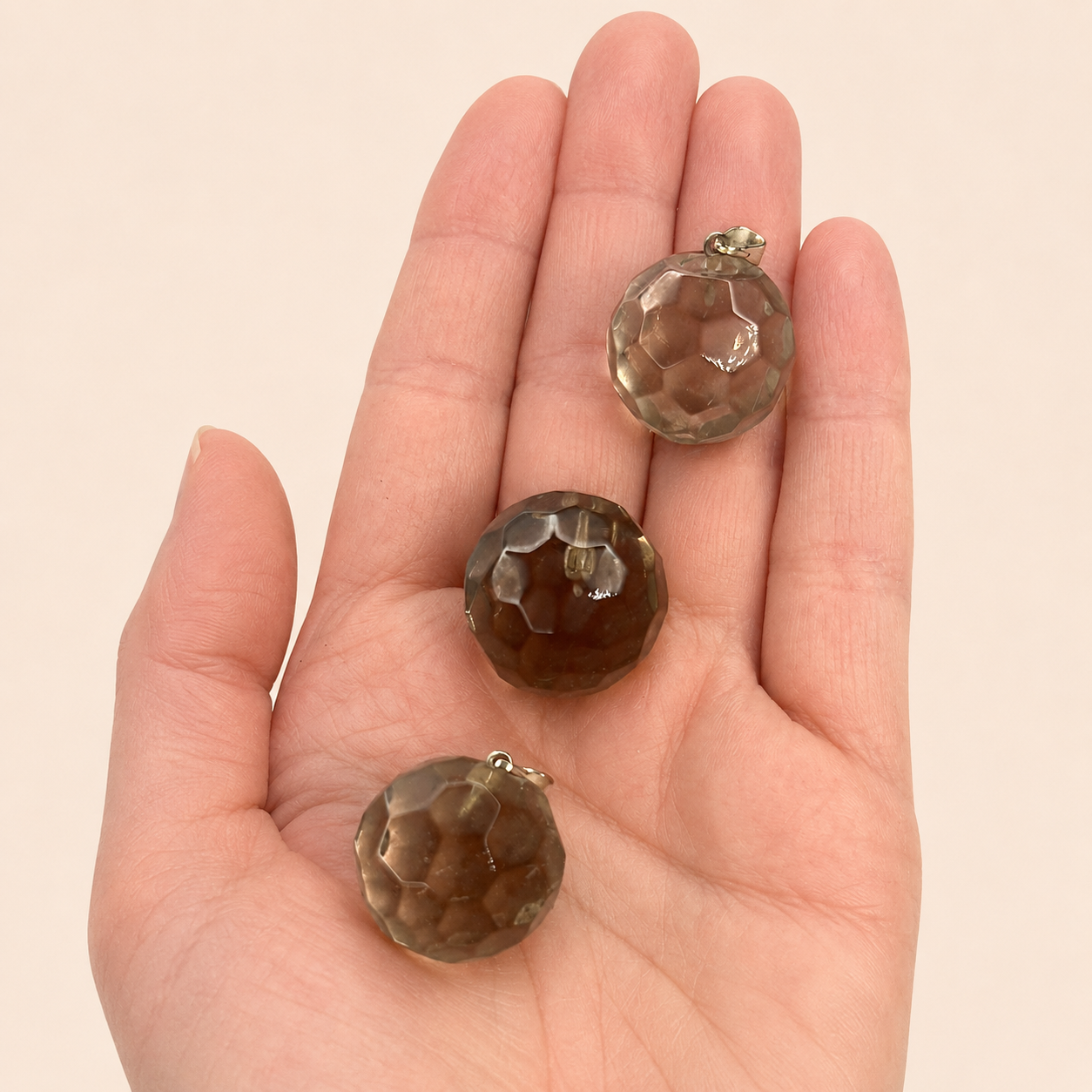 Three faceted brown smoky quartz beads with silver-toned metal loops, displayed on a person's palm against a light beige background.