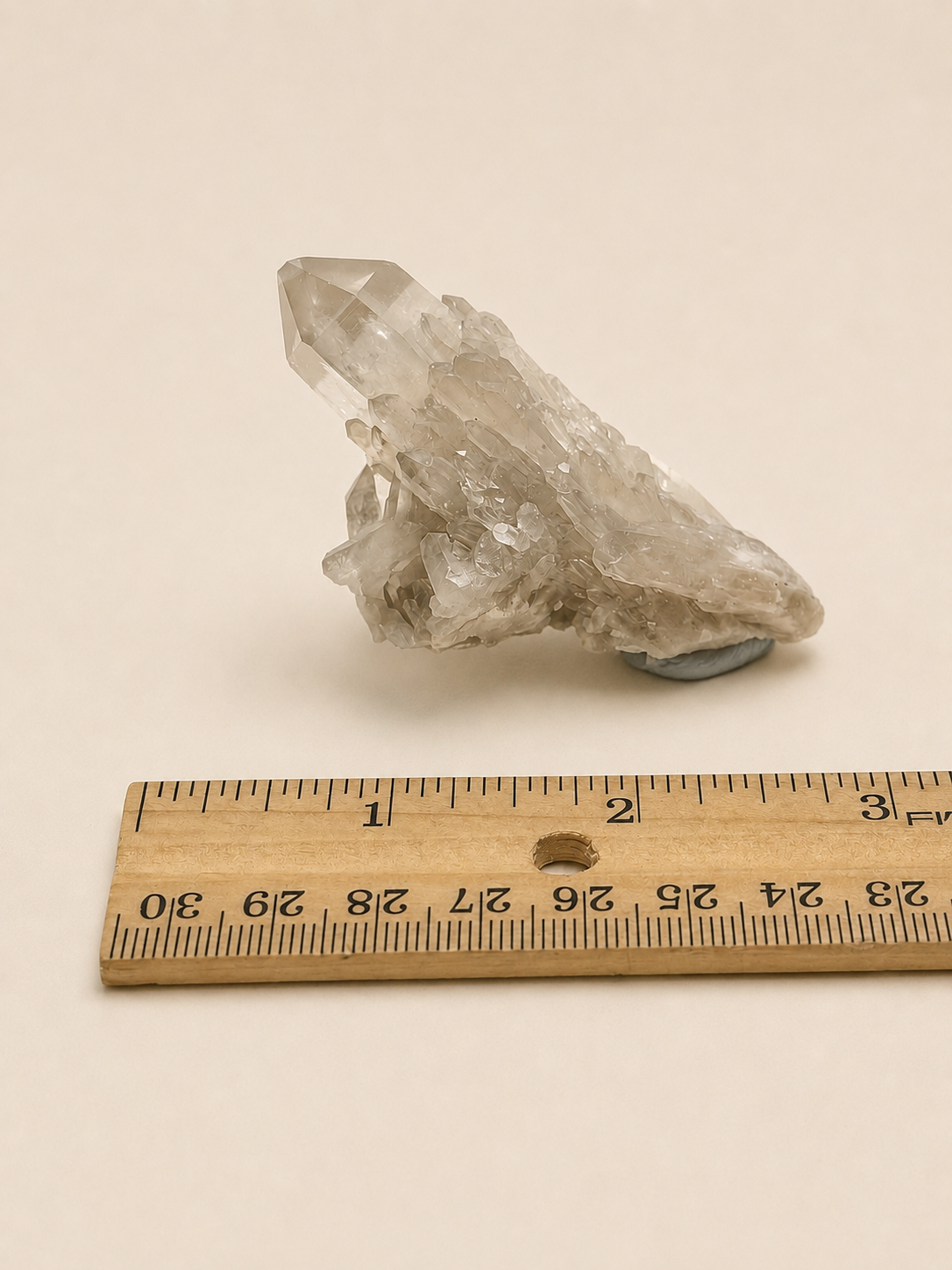 A raw clear quartz crystal cluster with a natural matrix, shown next to a wooden ruler for scale on a warm off-white background.