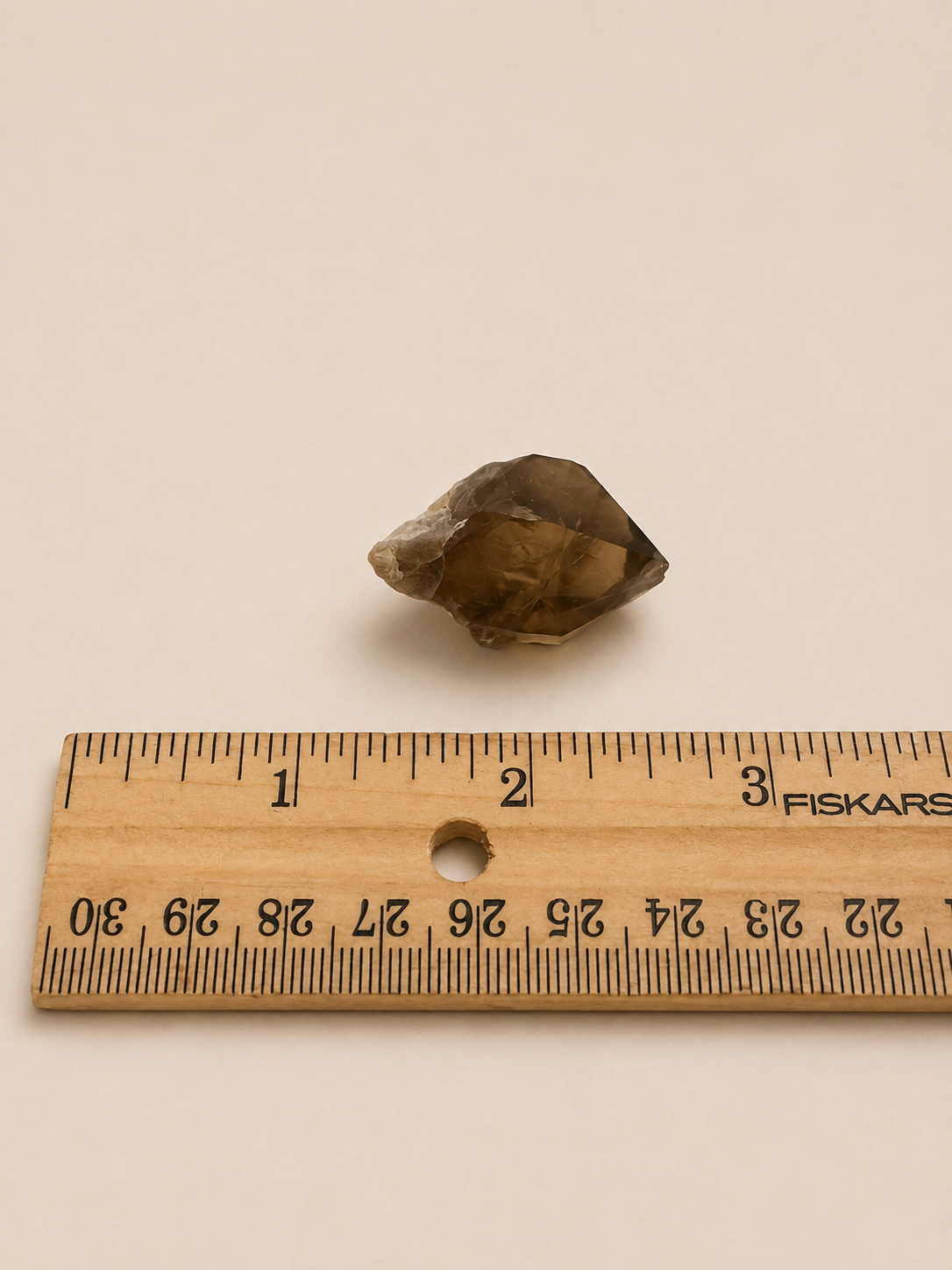 A small rough brown gemstone or crystal placed above a wooden ruler on a plain light beige background. The ruler shows measurements in centimeters and includes the visible brand name FISKARS.
