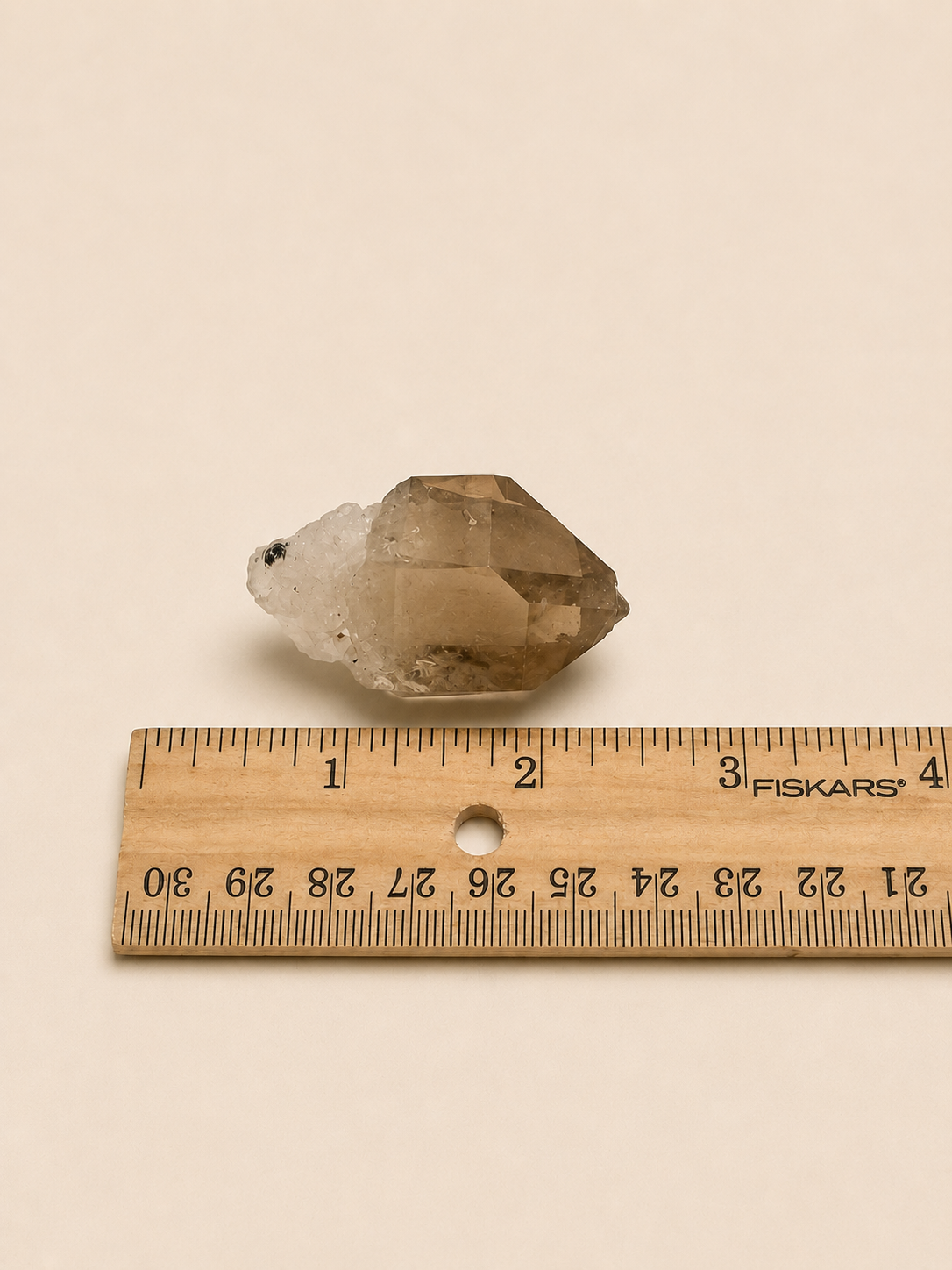 A small translucent smoky crystal point placed above a wooden ruler on a plain beige background. The ruler shows measurements in centimeters and includes the visible brand name FISKARS.