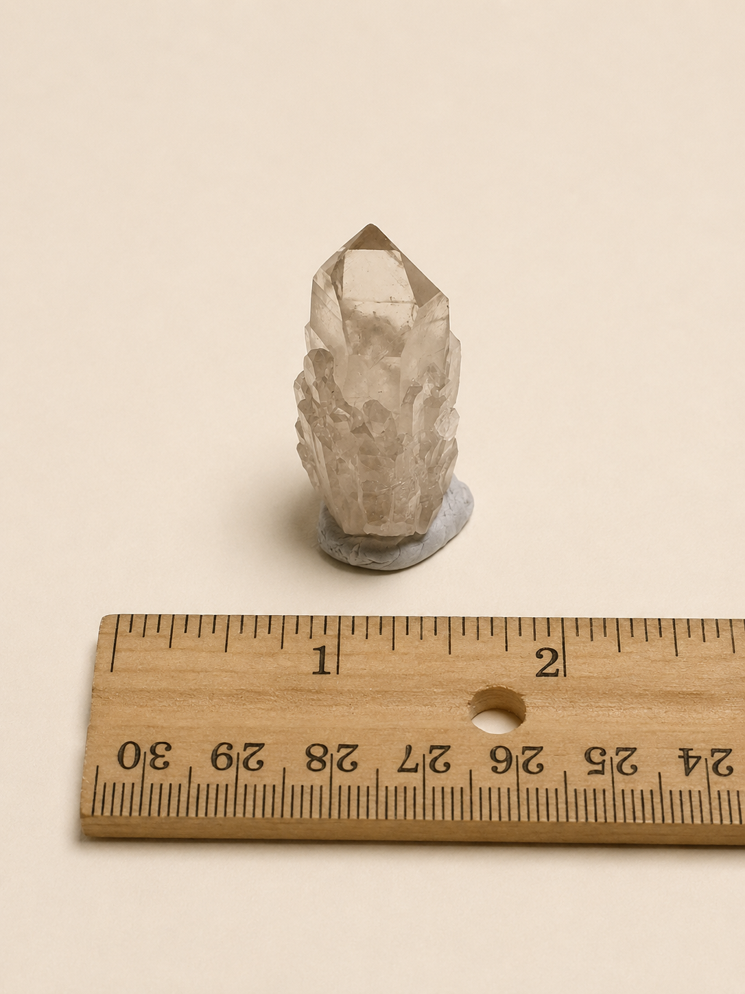 A small clear quartz crystal cluster on a gray base, shown next to a wooden ruler for scale on a warm beige background.