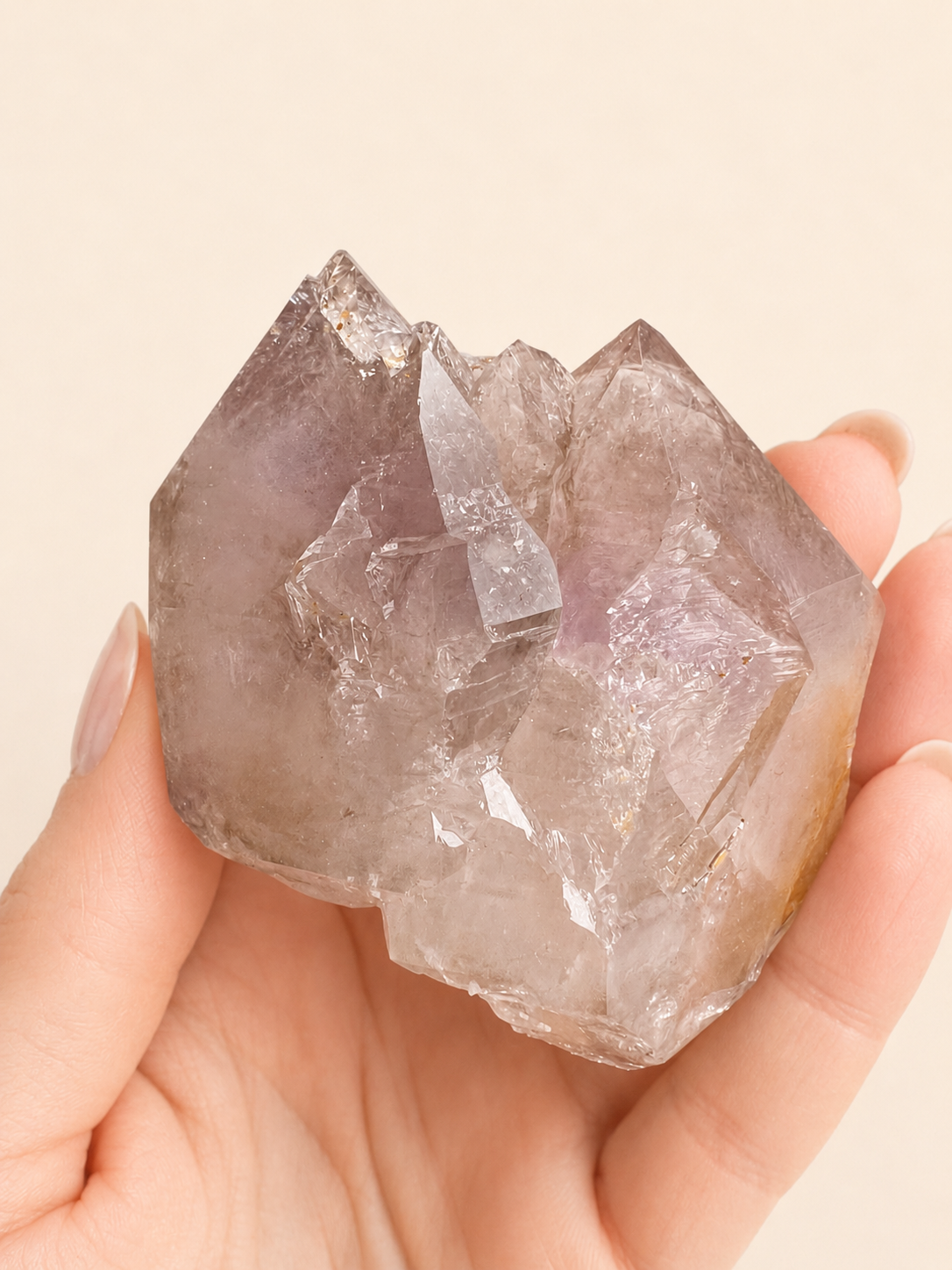 A hand holding a raw, rough crystal cluster with sharp facets and a translucent lavender-purple hue against a solid light beige background.