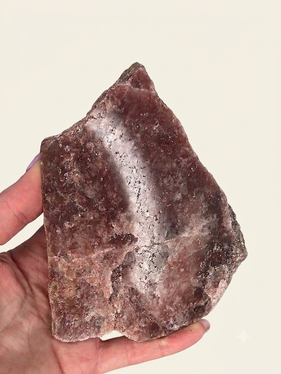 A hand holding a rough, irregularly shaped piece of rose quartz crystal, showing its natural texture and purplish-brown color against a plain light beige background.