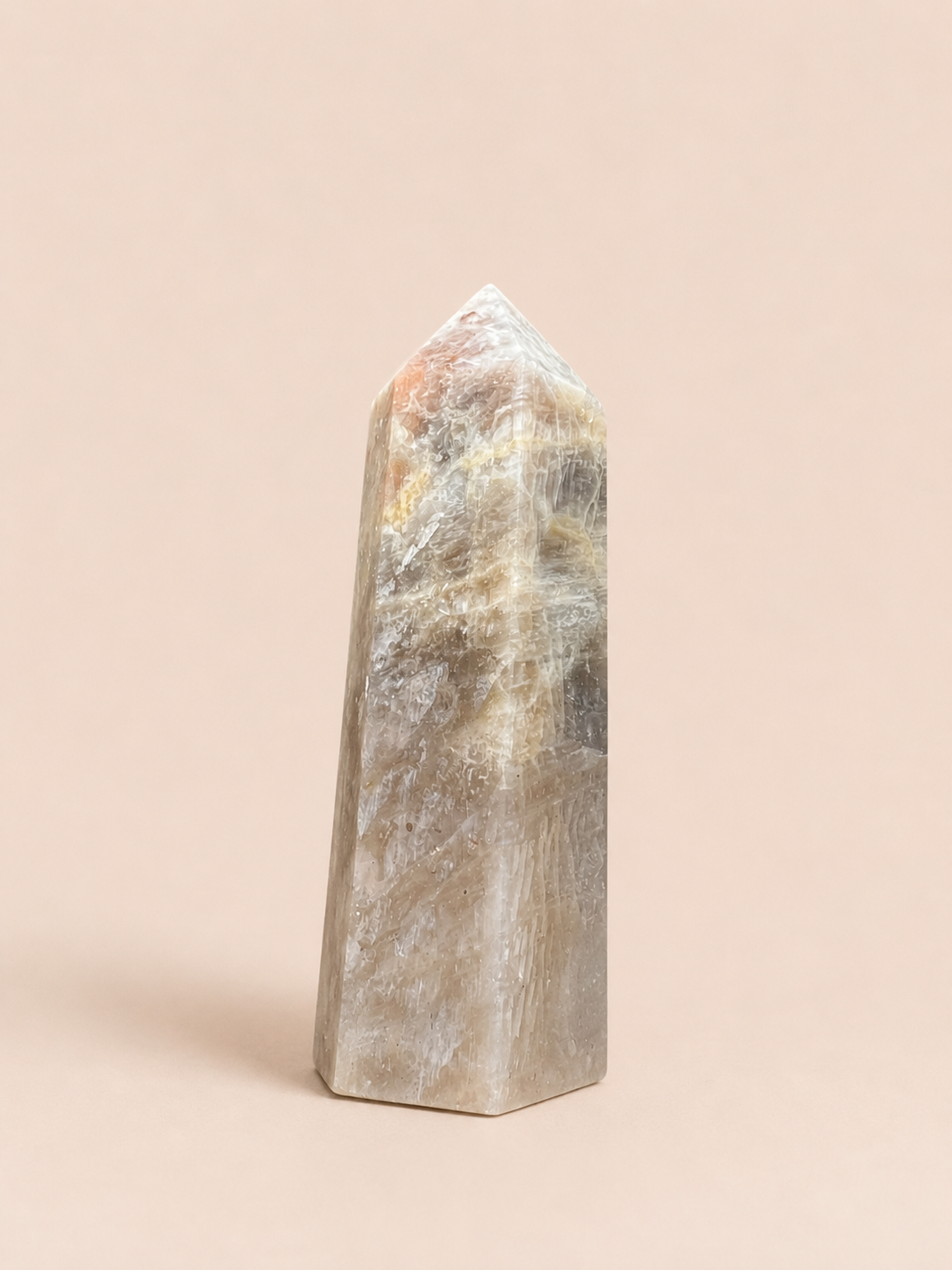 A polished natural crystal tower with a pointed apex, featuring a mix of gray, beige, and subtle orange inclusions, standing upright against a solid light beige background.