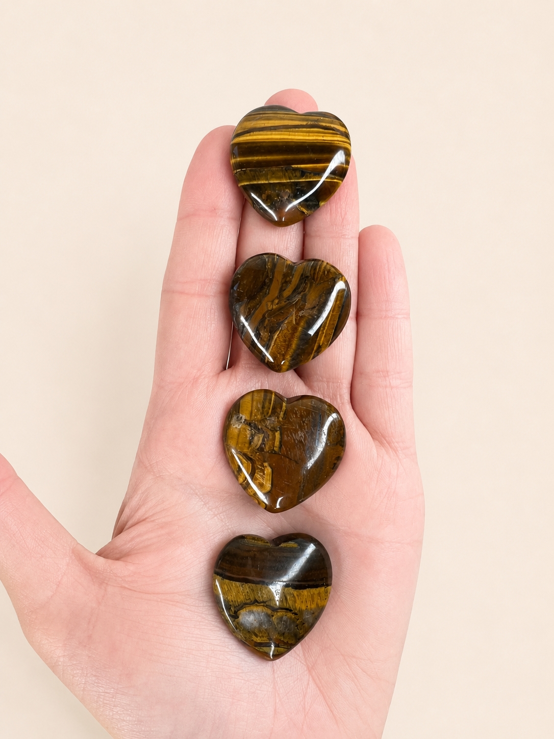 Four polished heart-shaped tiger's eye stones held vertically in a person's hand against a plain beige background.