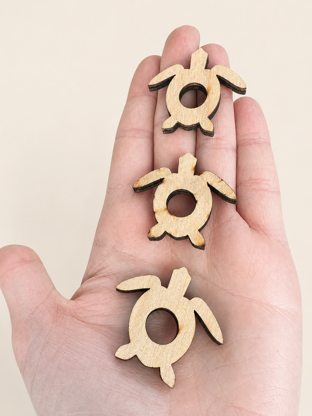 Three small laser-cut wooden turtle charms with a natural light wood finish and dark edge trim, held vertically in a person's hand against a plain beige background.