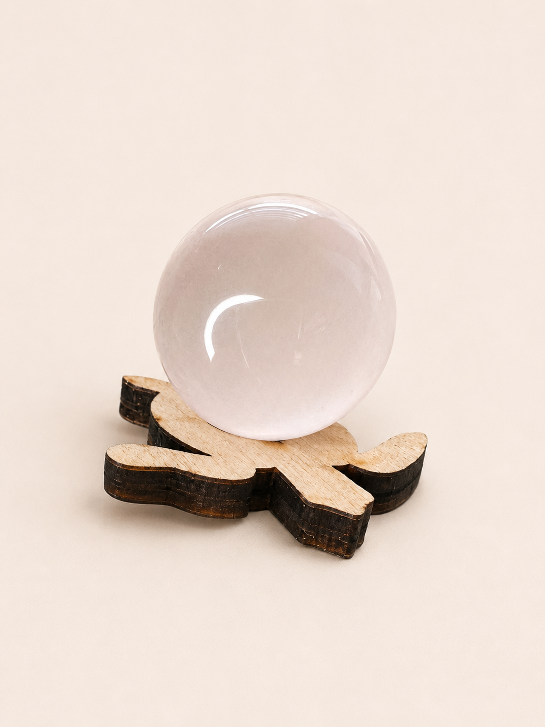 A smooth, translucent pale pink crystal sphere resting on a decorative wooden stand with dark and light wood sections, set against a solid beige background.