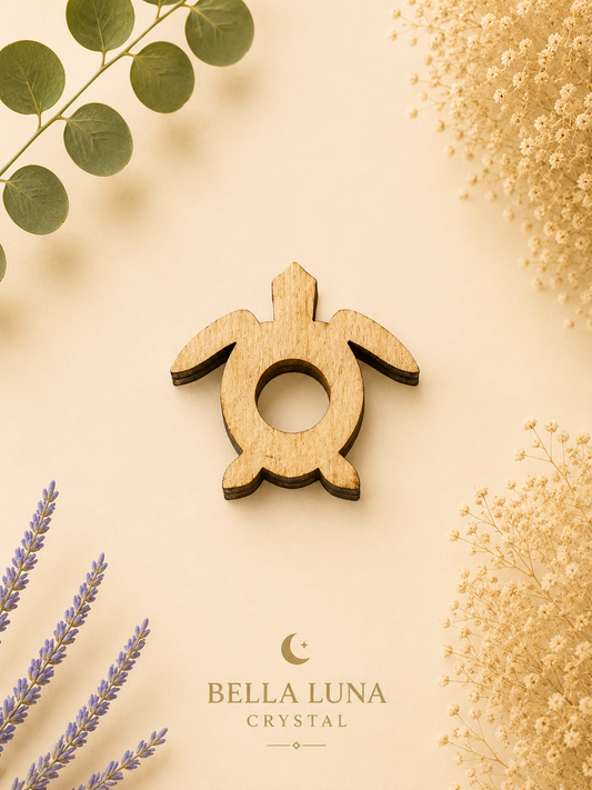 A wooden turtle-shaped crystal pendant from Bella Luna Crystal is displayed on a warm beige background with decorative botanical elements including eucalyptus leaves, lavender sprigs, and dried flowers.