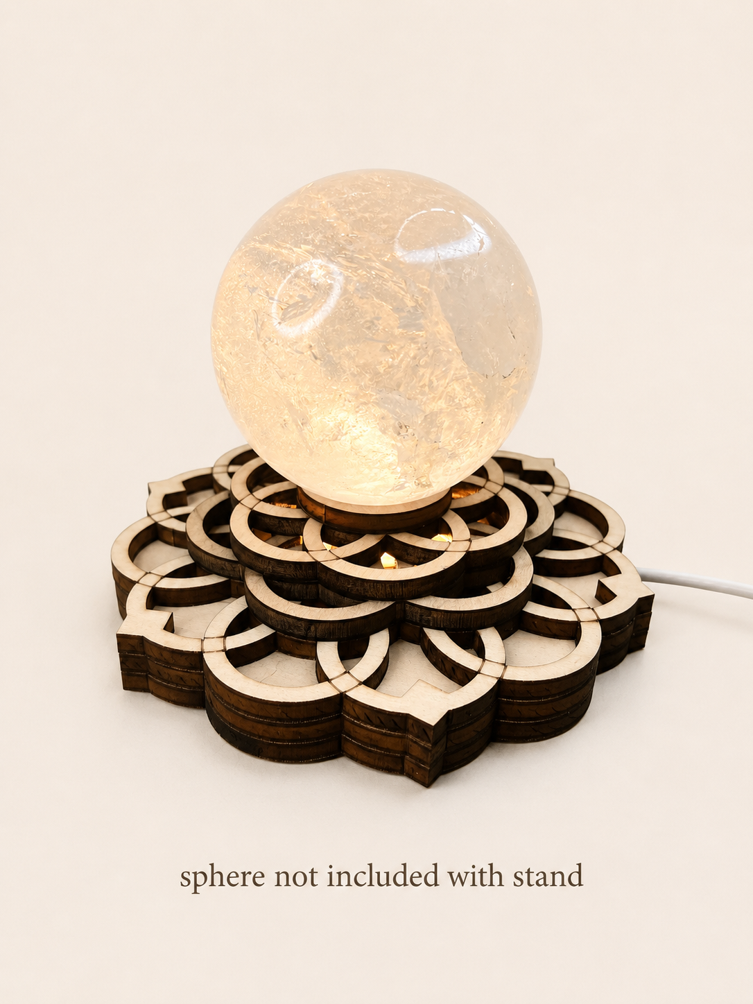 A decorative wooden stand with interlocking laser-cut circular patterns, supporting a clear glowing crystal sphere. The text 'sphere not included with stand' appears below, indicating the stand is the sold item.