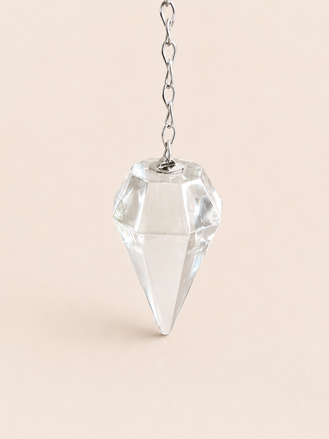 A faceted clear crystal cone pendant hanging from a silver chain against a solid pale peach background.
