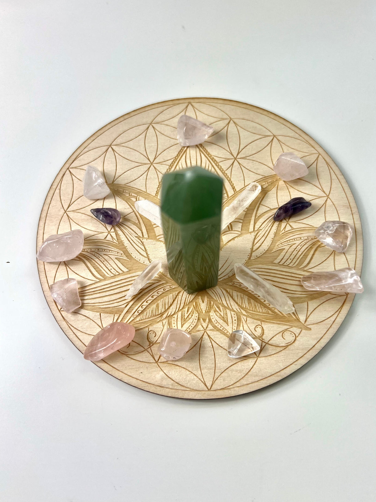 A green aventurine crystal pillar candle displayed upright in the center of a circular wooden holder engraved with a geometric floral pattern. The holder is styled with various raw crystal points, including pink rose quartz, clear quartz, and a purple amethyst, arranged symmetrically on a plain white background.